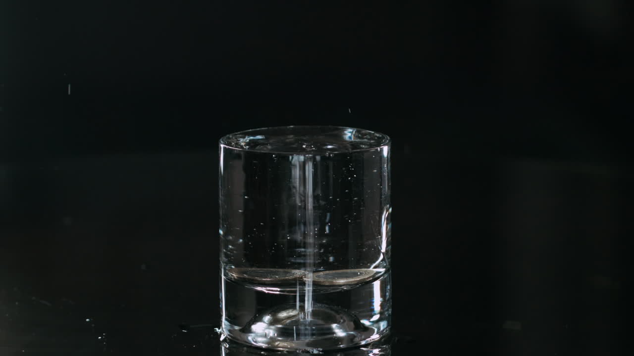떨어지는 금속성 토큰 동전을 물로 가득 찬 유리에 닫고, 극단적인 느린 동작으로 고립된 검은색 배경 주위에 액체를 뿌립니다.