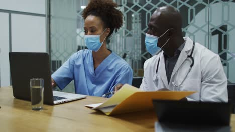 diverse female and male doctors wearing face masks using laptop and talking in office