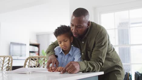Happy-african-american-man-and-his-son-sitting-at-table-and-reading-braille