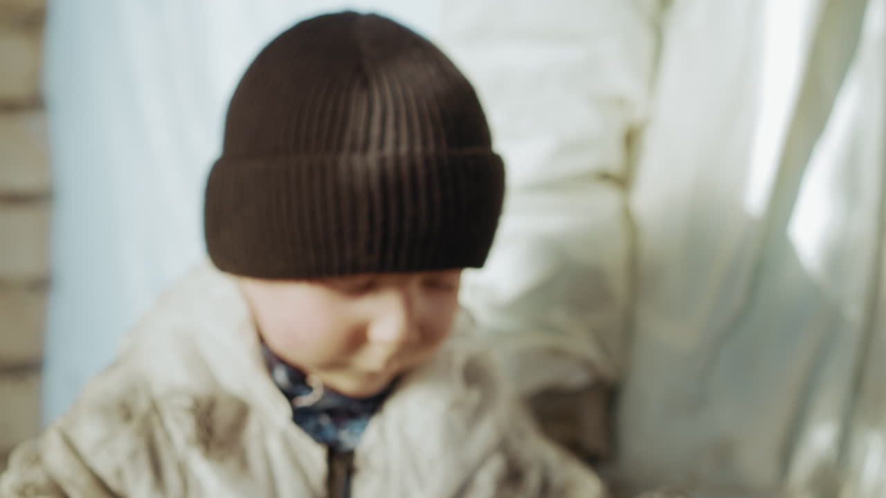 Child hand grips edge of oversized hazmat mask with green straps and metallic clasps, suggesting curiosity with creased dirty surface highlighted by natural indoor light in gritty setting