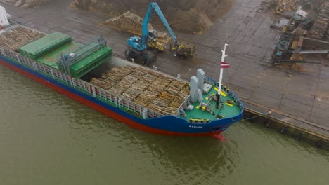 aerial establishing view of wood terminal crane loading timber into the cargo ship, port of liepaja , lumber log export, overcast day with fog and mist, birdseye drone moving backward