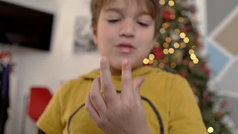 niño caucásico lamiendo azúcar glaseado de sus dedos, árbol de navidad en el fondo borroso 4k