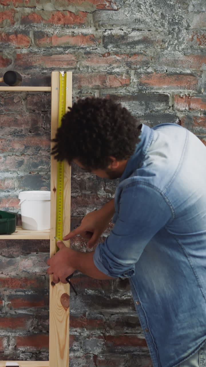 Black man measures wooden shelving unit with roller tape in living room backside view. African-American employee works in room prepared for renovation