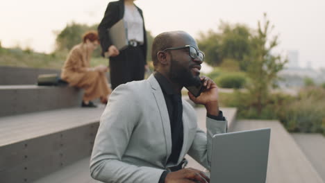 african american businessman talking on phone and using laptop outdoors