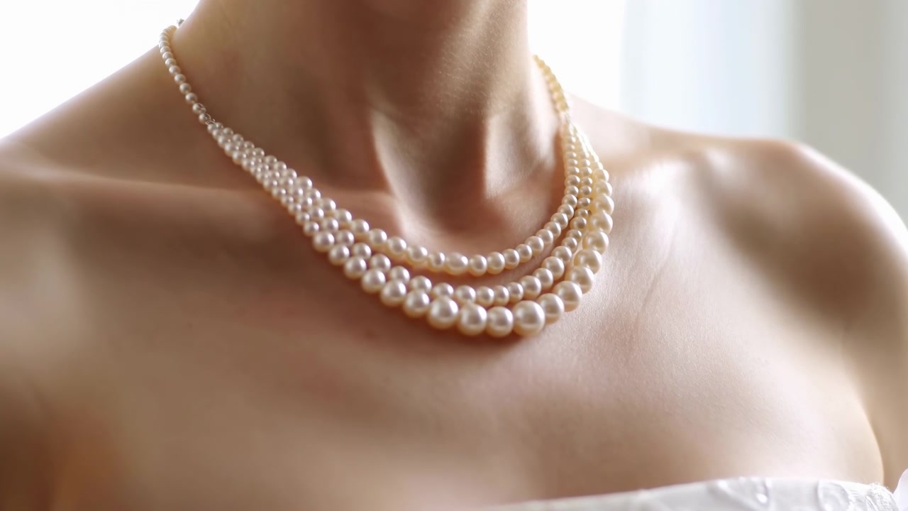 Primo piano in movimento di tre fili di perle a scalare che poggiano sul collo e sulle clavicole di una donna, con un sottile riposizionamento dovuto a un leggero cambio di postura sotto una morbida retroilluminazione dalla finestra, per un uso elegante nella moda