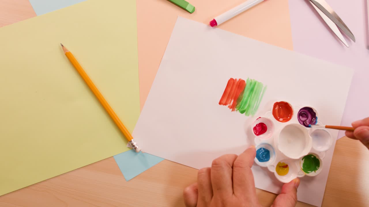 Hand uses paintbrush to apply vibrant colors on paper, overhead view, bright even lighting