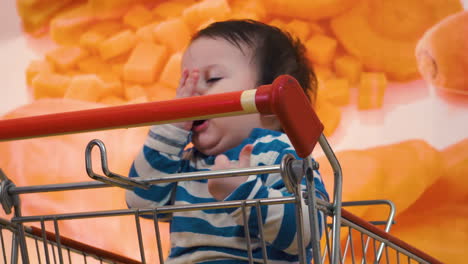 bebé cansado golpeándose ligeramente en la cara y lloriqueando mientras espera a la familia dentro de la tienda de comestibles sentado en el carro de la compra