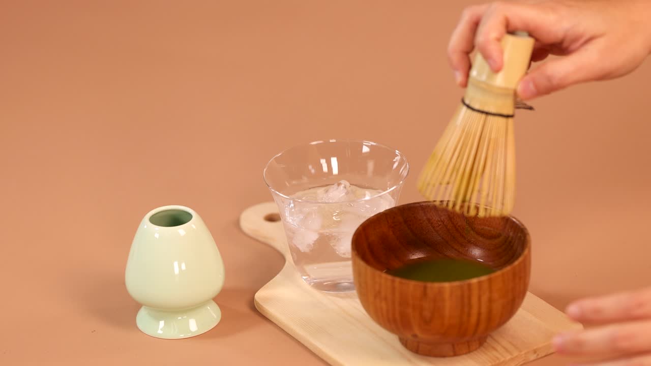 Hand uses bamboo whisk to mix matcha green tea in wooden bowl, minimal warm lighting