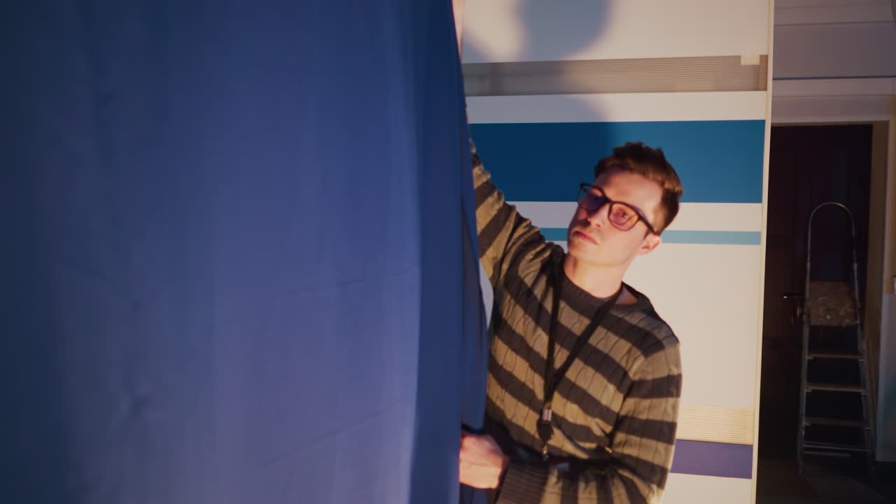 Male technician adjusts blue curtain on television studio set during behind scenes prep, floor spotlight illuminates striped backdrop while cables, ladder and rigging equipment lie ready for broadcast production