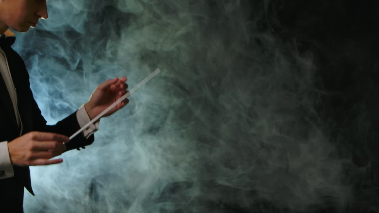 옷을 입은 교향악단 지휘자는 연기가 있는 검은색 배경에서 막대기의 움직임으로 음악가들을 지휘하고 있으며, 가시적인 몸짓으로 음악 공연을 지휘하고 지휘하고 있다.