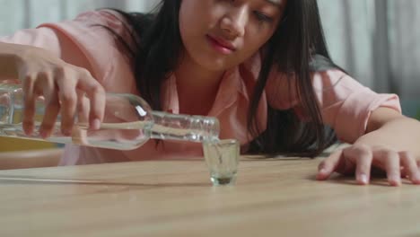 un primer plano de una mujer asiática borracha y deprimida con un teléfono inteligente en la mesa vertiendo vodka en un vaso antes de beber en casa.