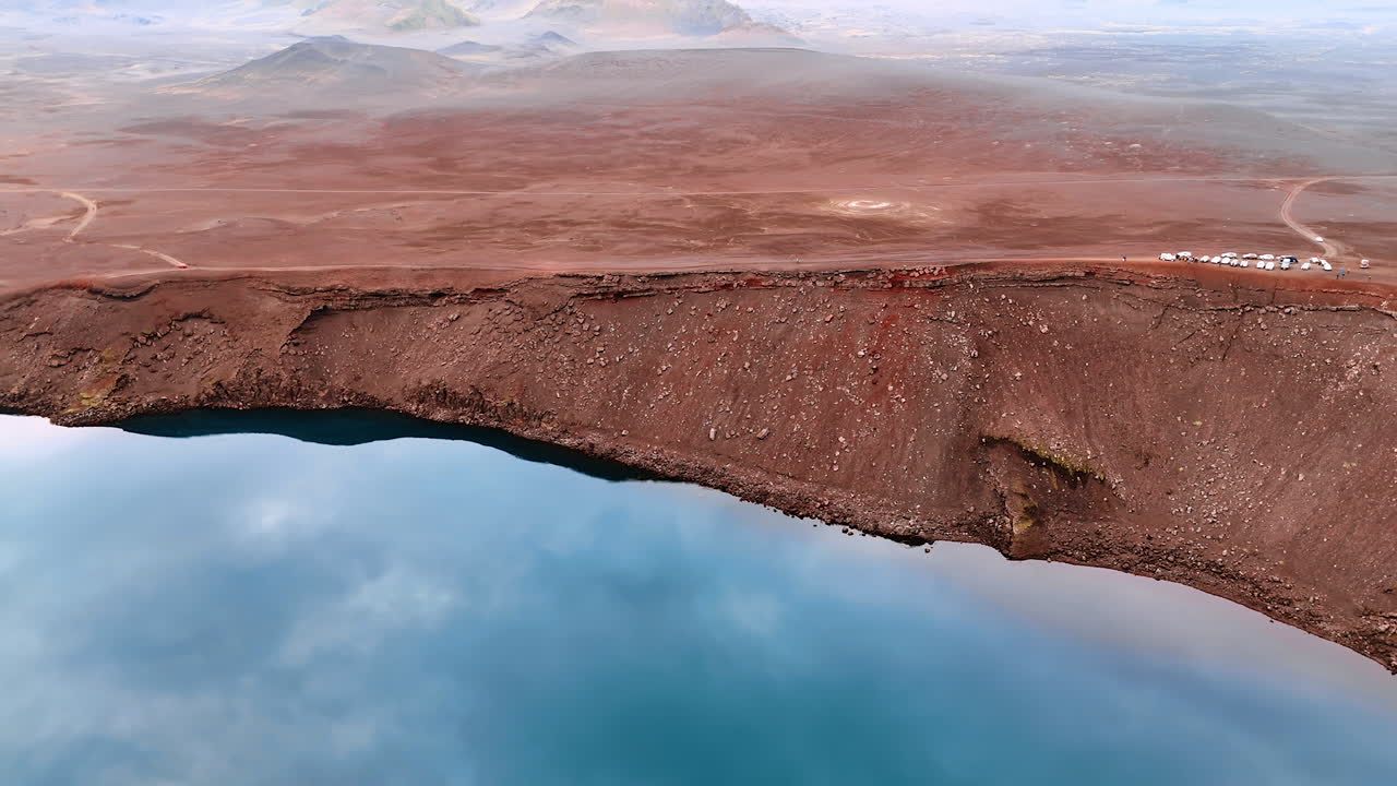 A group of tourist cars standing at the edge of a volcano crater. A lake with blue water is filling the crater. Aerial perspective on the landscape in Iceland.