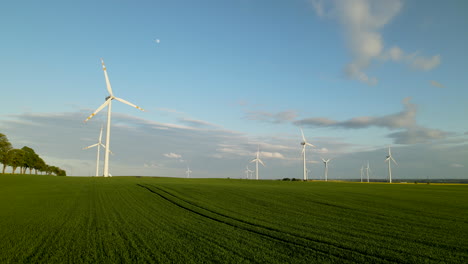 Amanecer-En-El-Parque-De-Turbinas-Eólicas-Con-Campo-Verde-Y-Luna-En-El-Cielo,-Antena