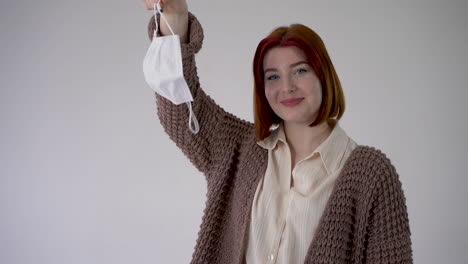 End-of-the-COVID-19-quarantine.-Young-cheerful-woman-takes-off-medical-face-mask.-Copy-space-and-white-background.-Confident-female-looking-at-camera.