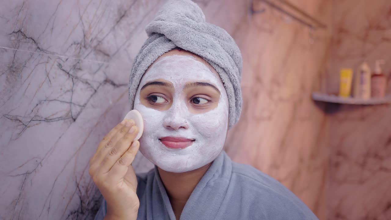 Close up shot of a young woman gently removing face pack with a cotton pad while looking into the bathroom mirror, showcasing daily skincare routine and self-care