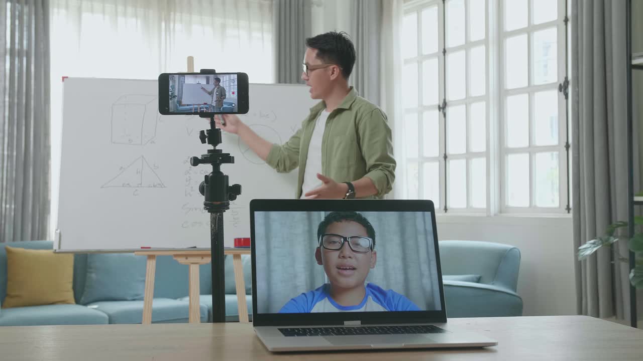 Asian Male Teacher With Glasses Shoots Video By Smartphone Camera And Having Video Call On Laptop While Teaching Math At Home