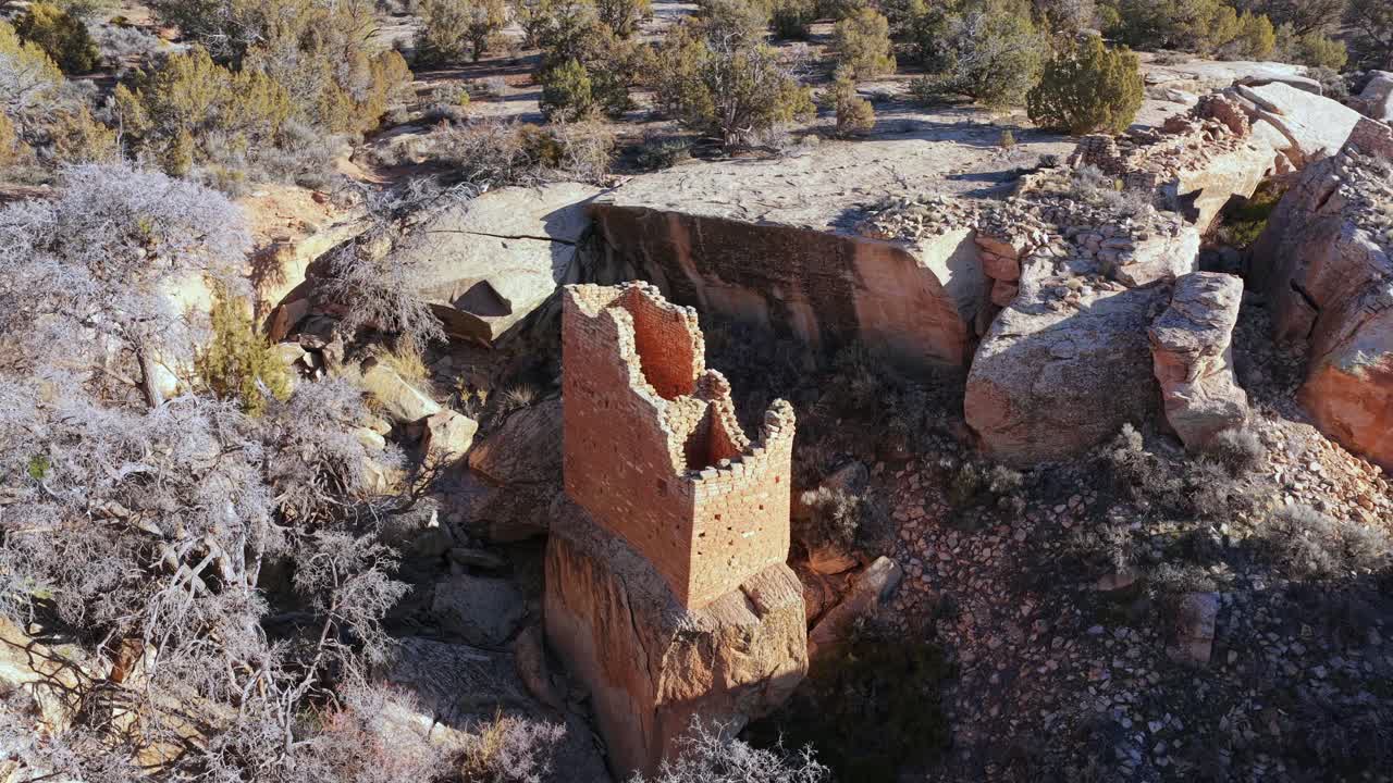 Un'alta rovina di pietra rettangolare si erge su un piedistallo di arenaria. La telecamera si allontana e si inclina verso il basso per rivelare la precaria muratura sullo sfondo della bellezza selvaggia di un canyon desertico occidentale.