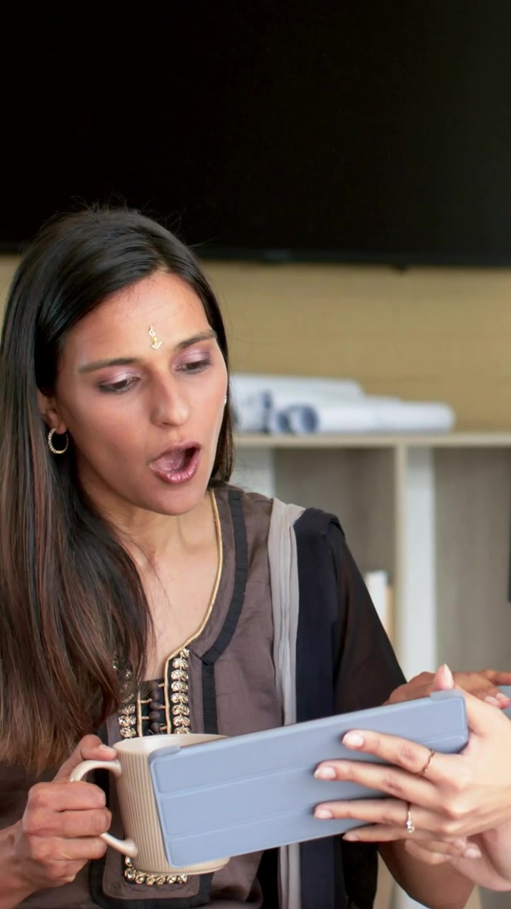 Video verticale: Indiano seduto con maglietta marrone che reagisce mentre un collega mostra un tablet blu in ufficio con una tazza.