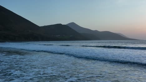 rossbeigh beach, county kerry, ireland, march 2022. drone tracks left in slow motion as gentle waves crash with whitewash rolling up along beach draining water through rocks at the dusk blue hour.