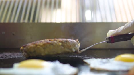 volteando la hamburguesa en una parrilla con humo saliendo y huevos alrededor de la hamburguesa a cámara lenta