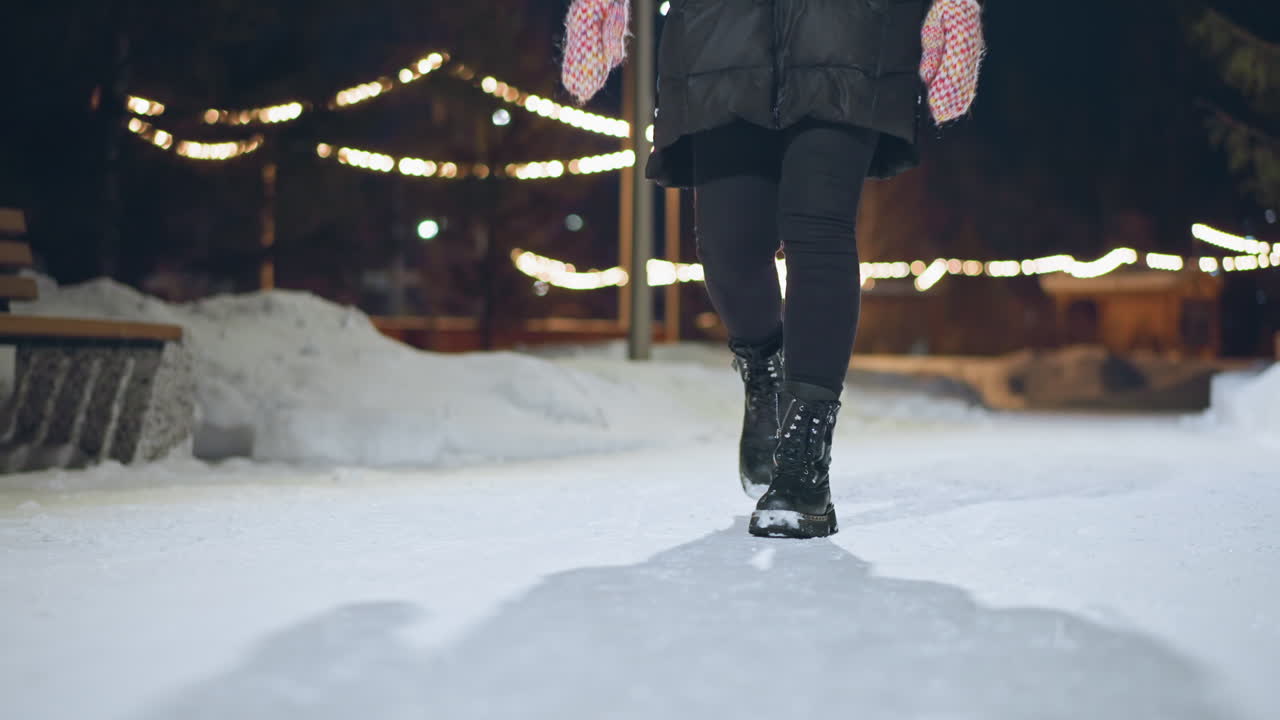 Persona con botas de invierno y ropa de abrigo caminando por un sendero nevado de noche, con luces festivas brillantes al fondo y una sombra que se extiende sobre la nieve, capturando el ambiente acogedor de un paseo en una tarde fría.