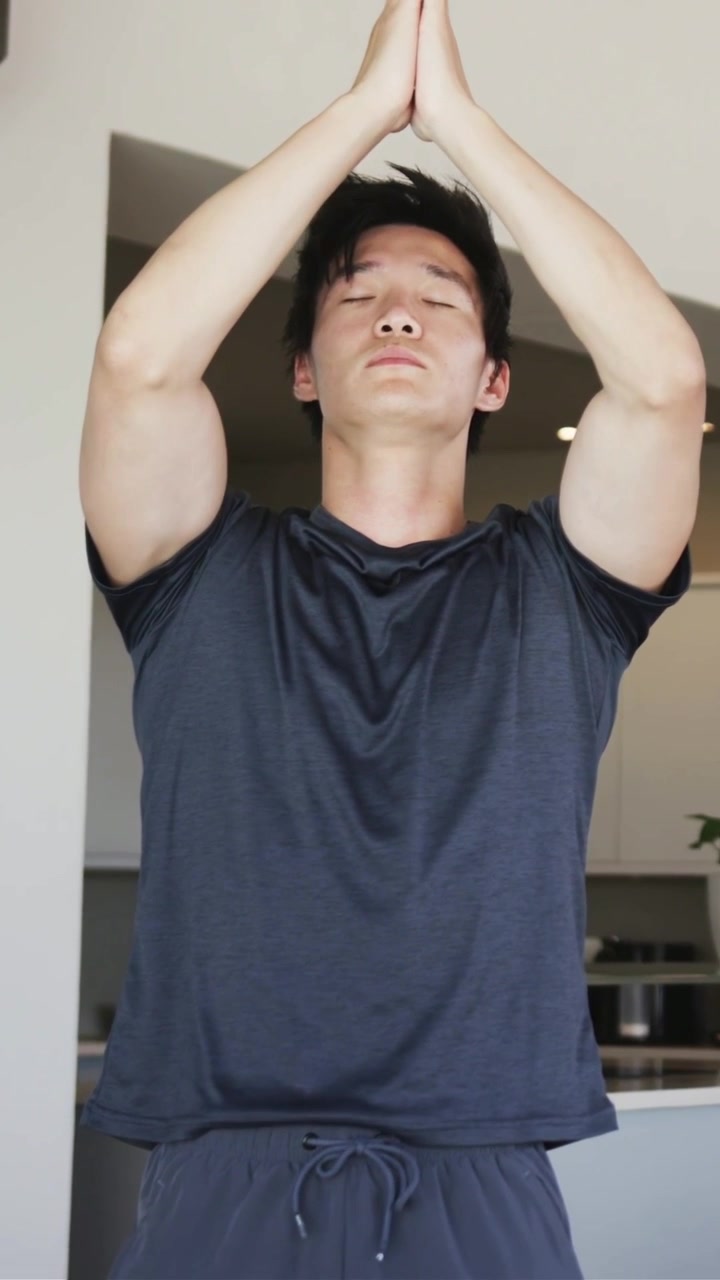 Video vertical: Hombre asiático con camiseta levantando los brazos, llevando las manos al pecho y orando en la cocina.
