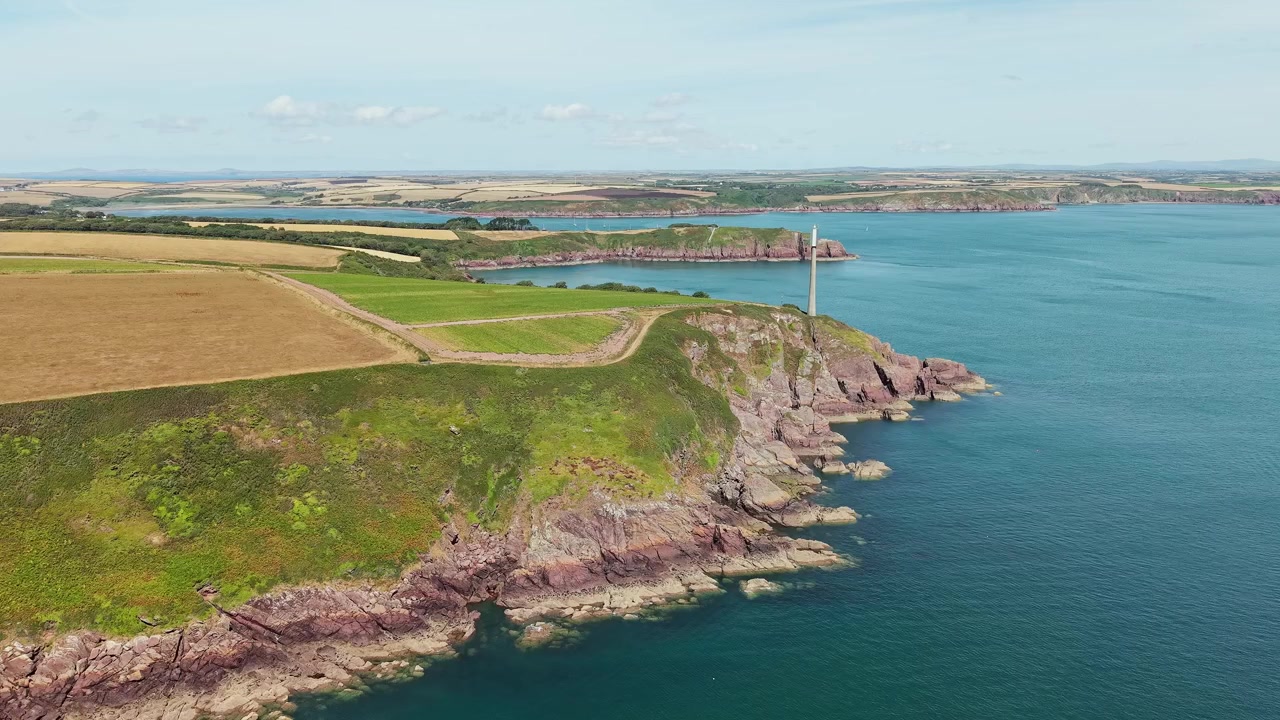 Drammatico promontorio roccioso con faro di navigazione che si affaccia sulle acque costiere, pascoli verdi e campi coltivati sotto il sole splendente dell'estate, Galles del Sud