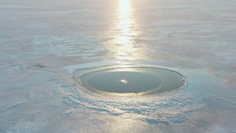 Tilt-Up-Drone-Shot-of-Bubbling-Ice-Hole-Revealing-Sunset-Over-Lake