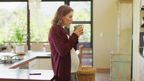 Mujer-Embarazada-Caucásica-Parada-En-La-Cocina,-Tocando-El-Vientre-Y-Bebiendo-Té