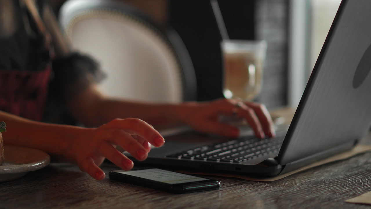 Cute young female adult with grin on phone while working on laptop computer at desk next to coffee cup with large window in background