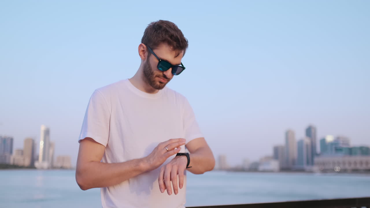 Happy handsome man in sunglasses and white t-shirt uses a watch watch looks and presses his finger on the screen standing on the waterfront in summer against the city and buildings