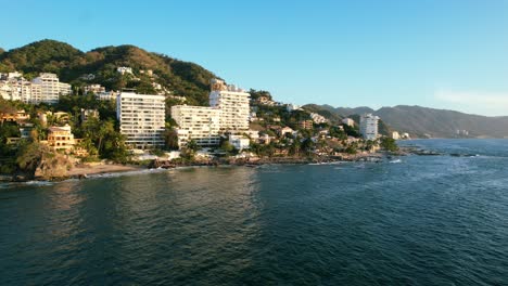 Hotels-and-resorts-on-oceanfront-mountain-hill-at-golden-hour-sunset-at-Los-Muertos-Beach-in-Puerto-Vallarta-Mexico,-aerial