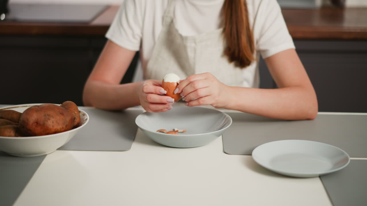 Woman peeling boiled egg over dish on kitchen table with scattered shell fragments inside plate, bowl of potatoes to left and empty plate to right