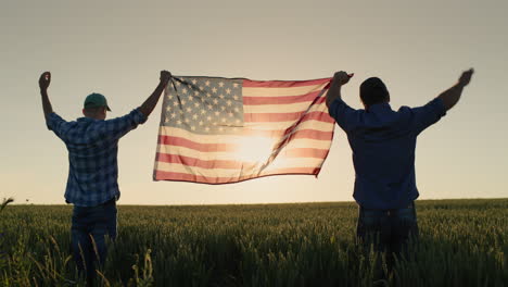 two men raise up the us flag against the backdrop of a wheat field at sunset