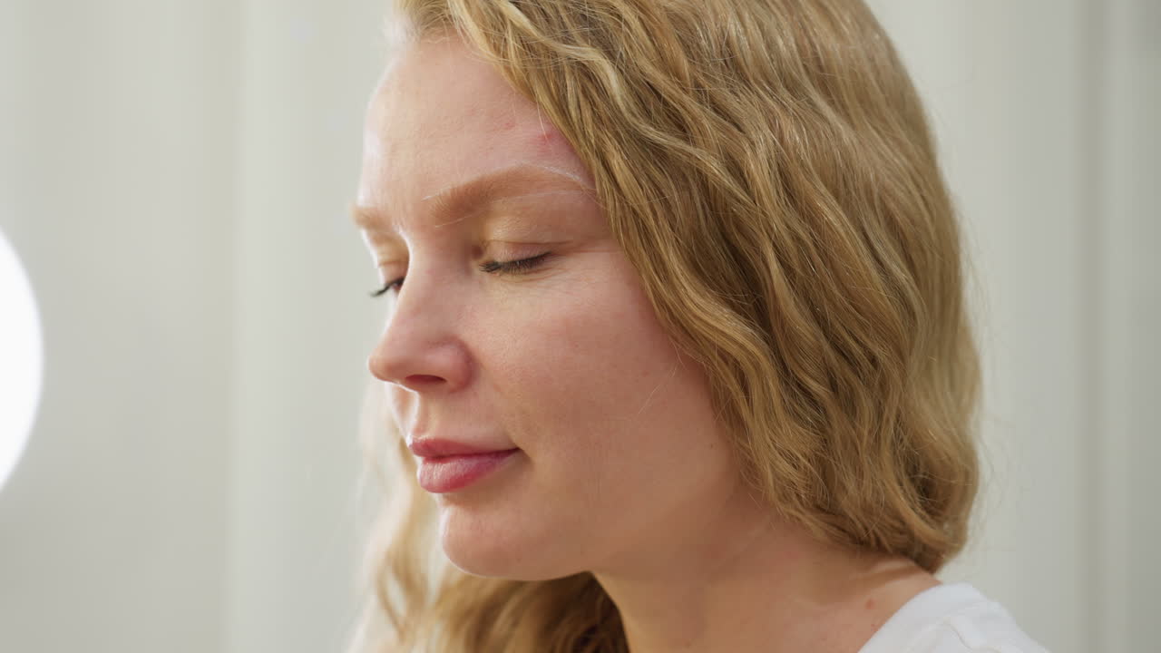Blonde-haired client with styled waves closely examines freshly shaped eyebrow in softly lit salon setting. Calm expression shows satisfaction and focus as she inspects cosmetic work done