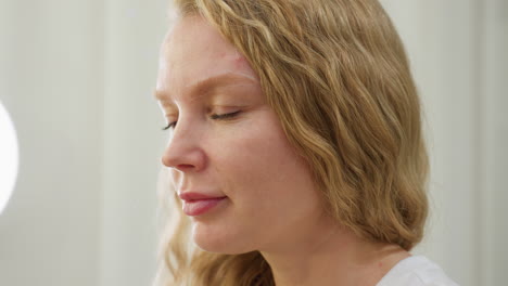 blonde-haired client with styled waves closely examines freshly shaped eyebrow in softly lit salon setting. calm expression shows satisfaction and focus as she inspects cosmetic work done