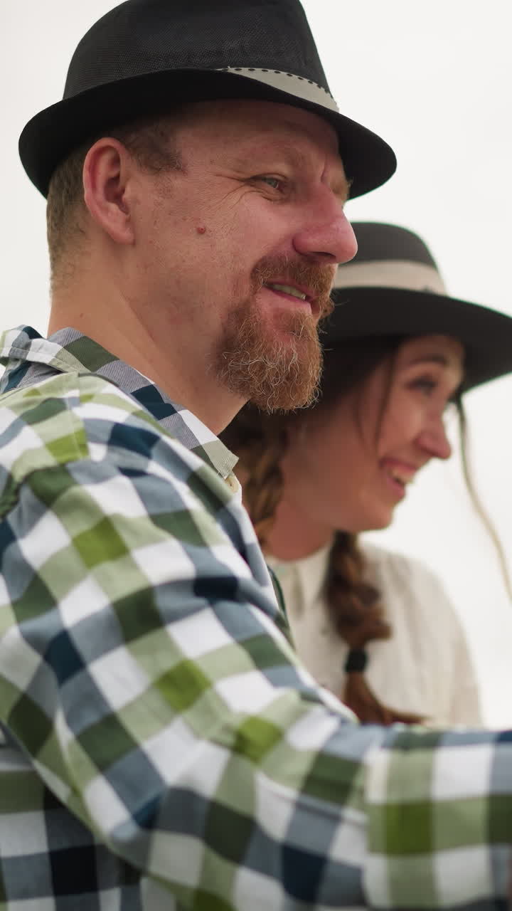 un artista feliz sosteniendo una paleta de pintura y trabajando en un gran lienzo al aire libre.sonríe mientras pinta, mientras que a su lado, una mujer con un vestido blanco y sombrero está de pie, observando alegremente su trabajo