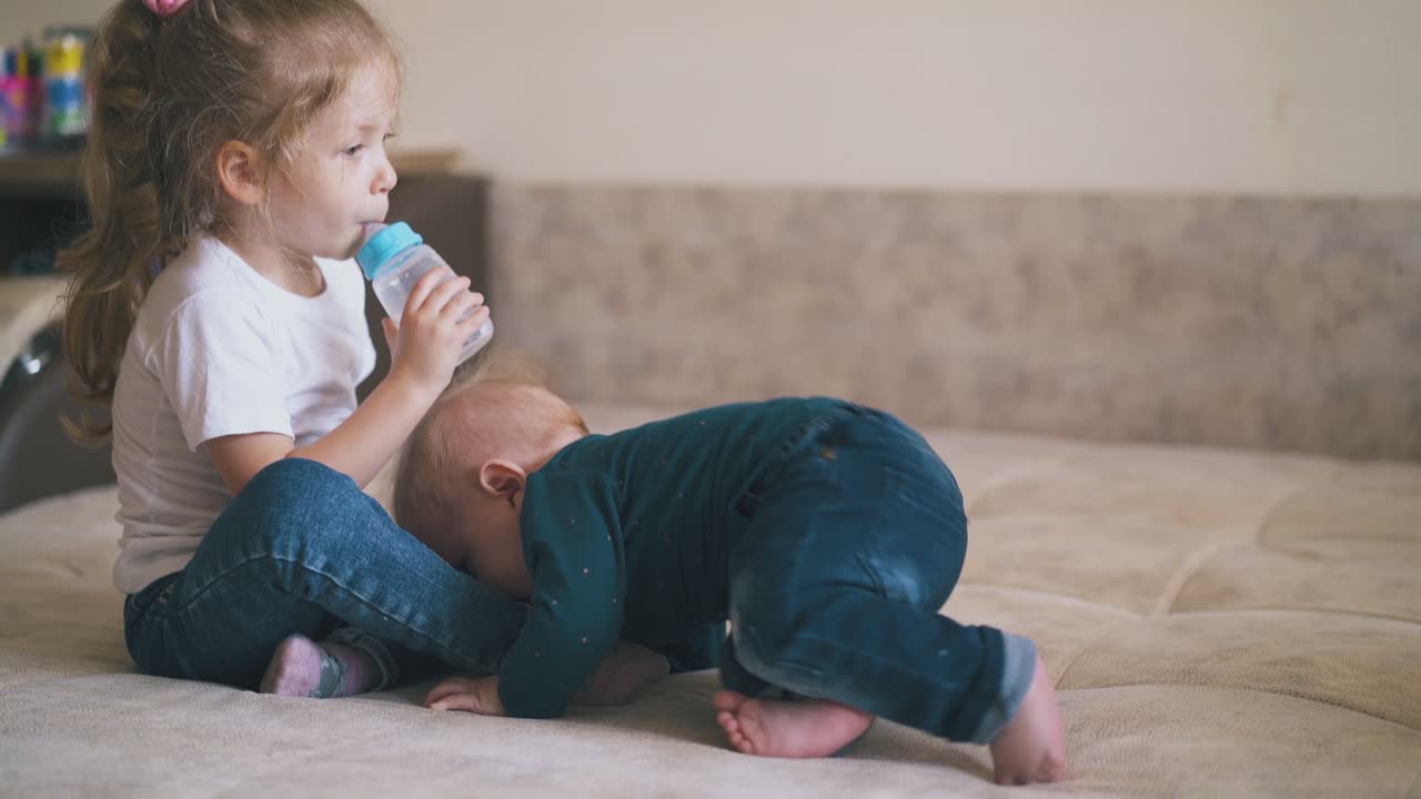 la chica chupa la tetina en la botella de agua y el hermano pequeño en la cama