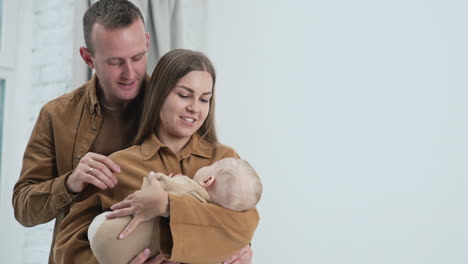 mother swaying her little infant child. loving husband stand behind stoking his wife and takes baby hand to kiss.