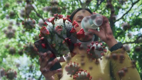 animation of covid 19 cells over woman in park using smartphone wearing face mask