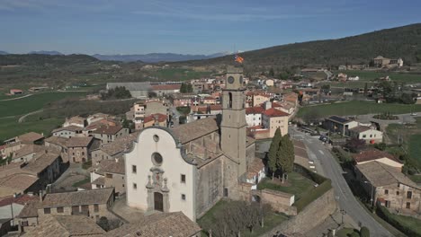 Vista-Aérea-De-Una-Iglesia-Típica-De-Un-Pueblo-De-Cataluña,-Cuyo-Campanario-Está-Coronado-Con-Una-Bandera-Independentista.