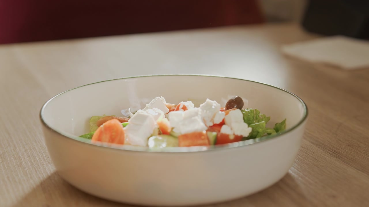 Close up of fresh salad in white bowl with green vegetables tomatoes and crumbled cheese on wooden table. Salad ingredients include lettuce cucumber slices cherry tomato pieces olives soft