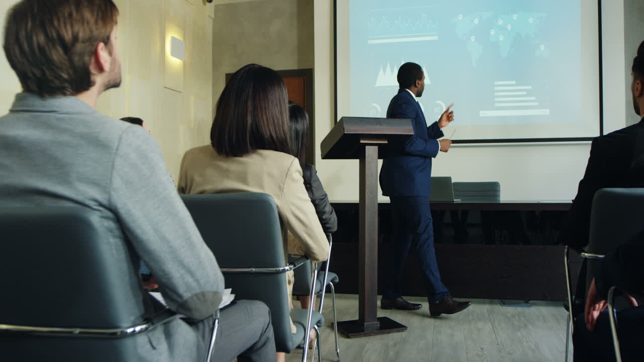 Camera zoom in on african american businessman talking on a podium in a conference room and showing some charts and graphics on the big screen