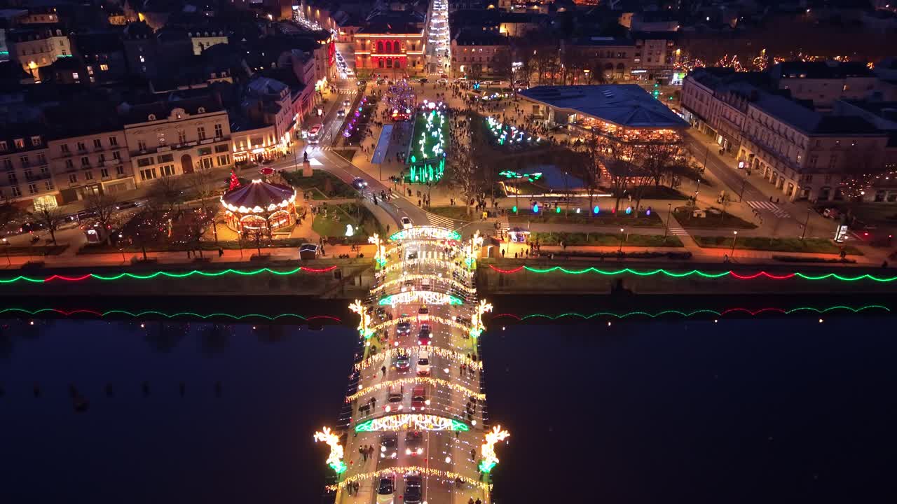 Drone advancing above Pont Aristide Briand in Laval, showing Christmas lights, crowds, traffic, reflections on the Mayenne, and the illuminated Place du 11 Novembre at dusk