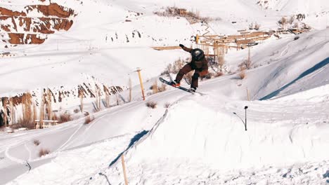 slow motion, drone shot, wide: pro snowboarder jumping in fresh snow, spraying snowflakes over sun. male freerider takes off in the air on his snowboard on sunny day on the mountain slopes