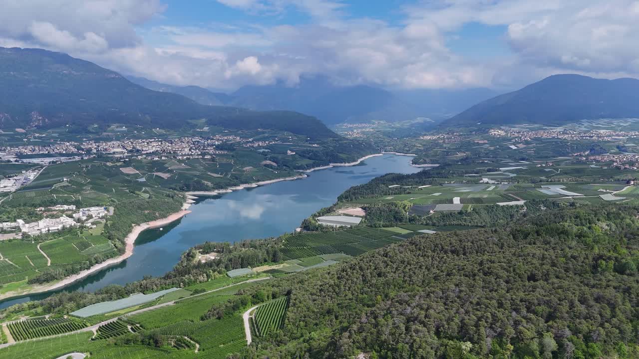 Drone video of Lake Santa Giustina, showcasing surrounding greenhouses, farmland, and a valley with mountains under a partly cloudy sky