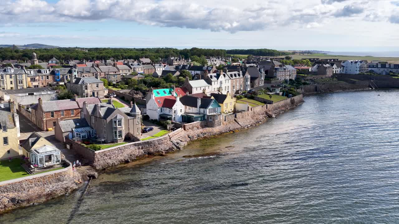 Drone glides above seaside village rooftops, revealing historic architecture, stone walls, and calm shoreline