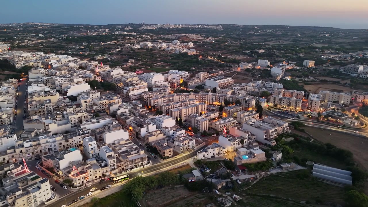 Paisagem urbana de Rabat, Malta com edifícios de pedra iluminados, telhados, estradas, tráfego e transporte público ao anoitecer em uma área residencial mediterrânea, vista aérea
