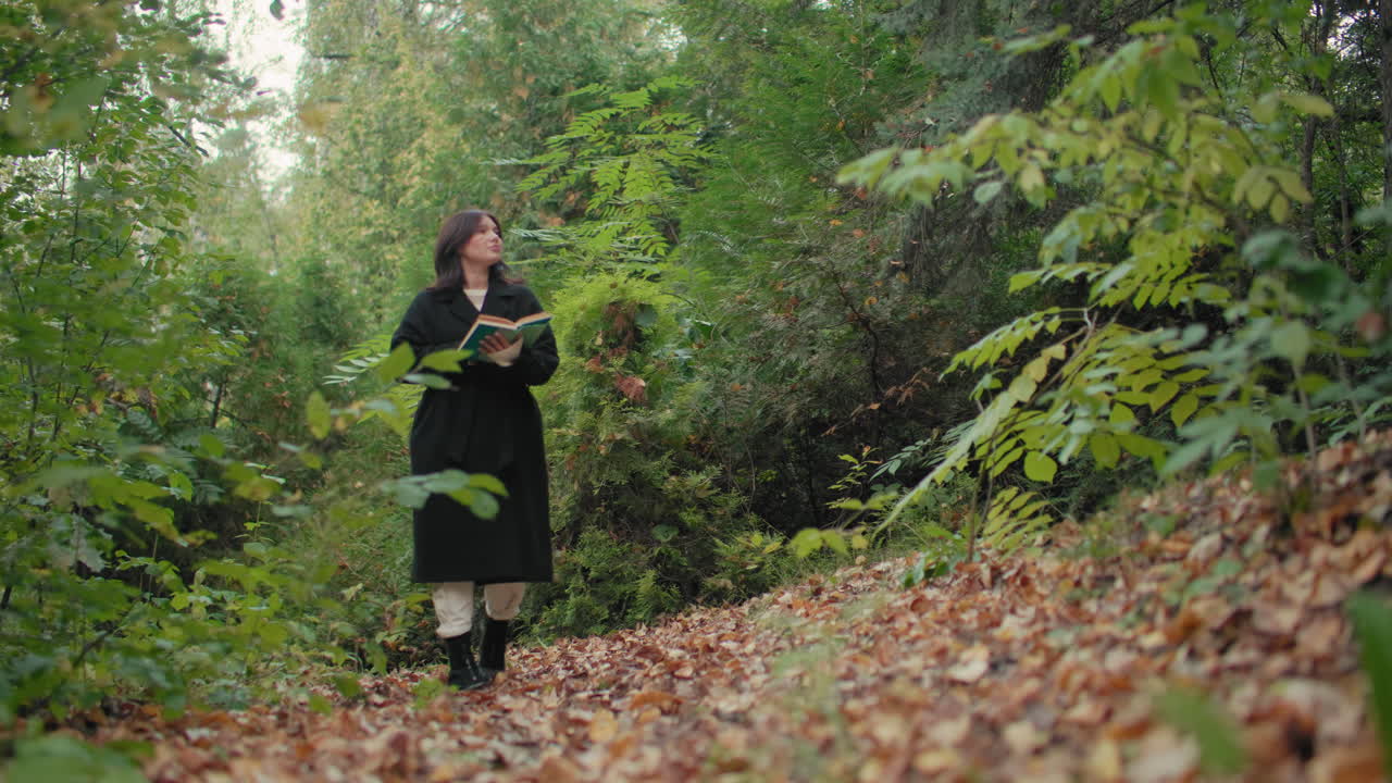 Una joven reflexiva pasea por el bosque con un libro, hojeando las páginas mientras disfruta de la naturaleza, un paseo tranquilo por un sendero frondoso, ambiente otoñal, curiosidad y evasión lectora en medio del follaje verde