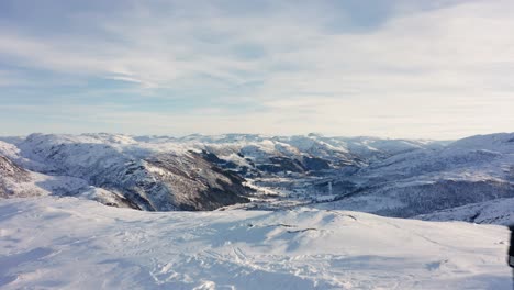 Toma-Aérea-Volando-Sobre-Un-Hombre-Parado-En-El-Pico-De-La-Montaña-Rodeado-De-Un-Paisaje-Nevado